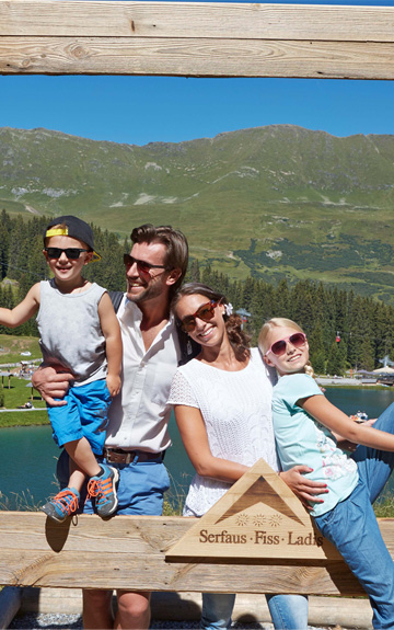 Eine Familie posiert in einem großen Holzrahmen vor einer Berg- und Seenlandschaft in Serfaus-Fiss-Ladis. Im Hintergrund sind grüne Almwiesen, Wälder, Berge und ein See mit Ausflugsgästen zu sehen.